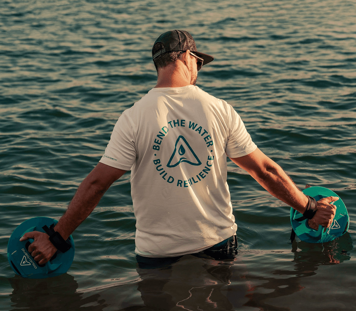 Man standing waist-deep in open water at sunset, arms outstretched, holding an Aqua-Bend disc in each hand
